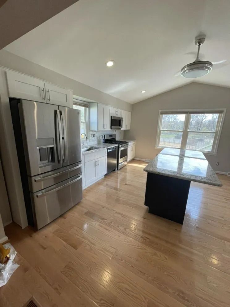 Kitchen with white cabinets, stainless steel appliances, and wooden floors. An island with a granite countertop is in the center.