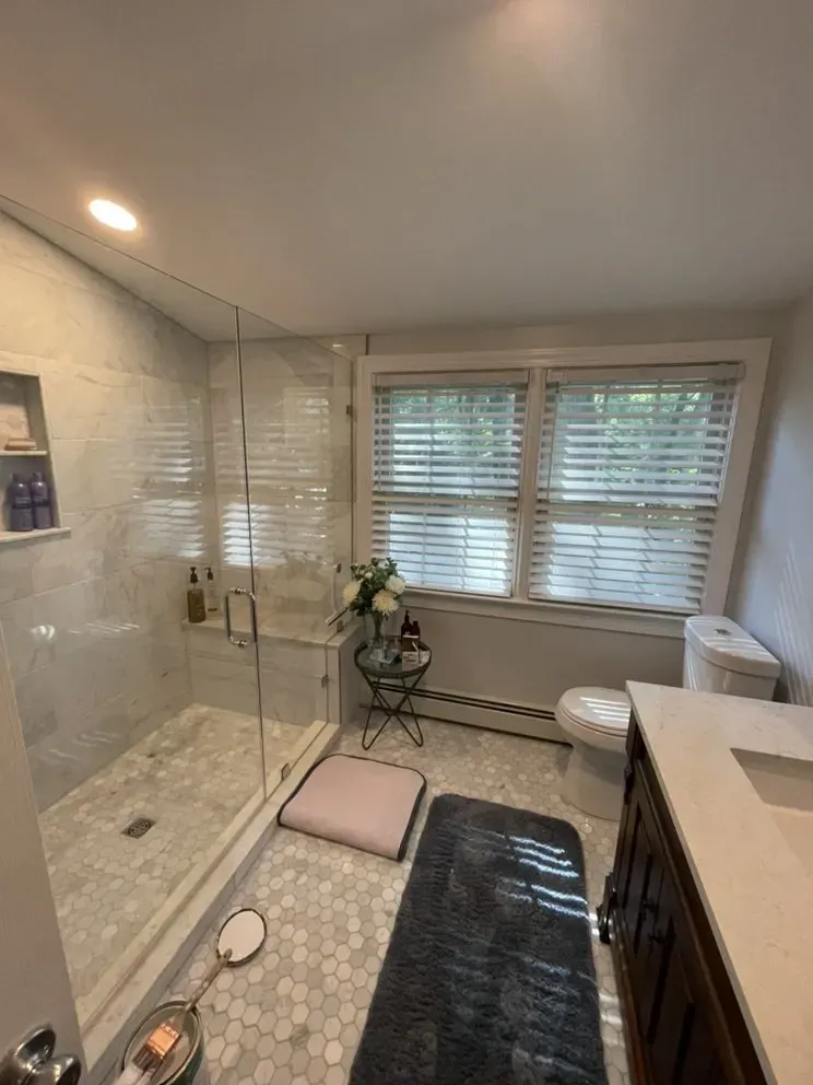 Bathroom with a tiled shower, toilet, vanity, and a window with blinds.