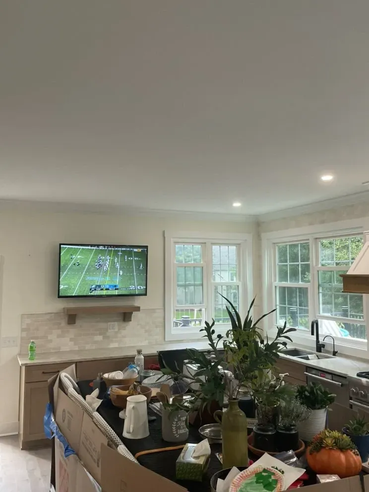 Kitchen with TV on wall, island with items, windows, and plants. TV shows a football game.