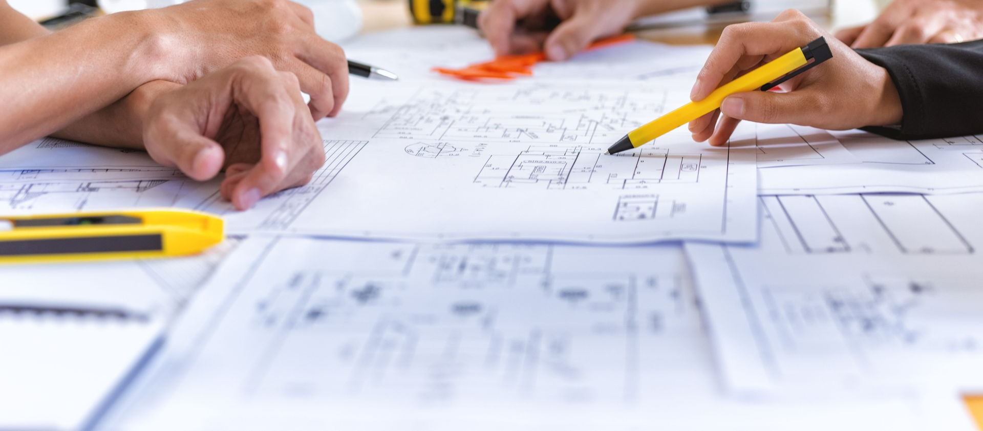 Hands of people discussing architectural blueprints. Yellow pen points to a drawing.