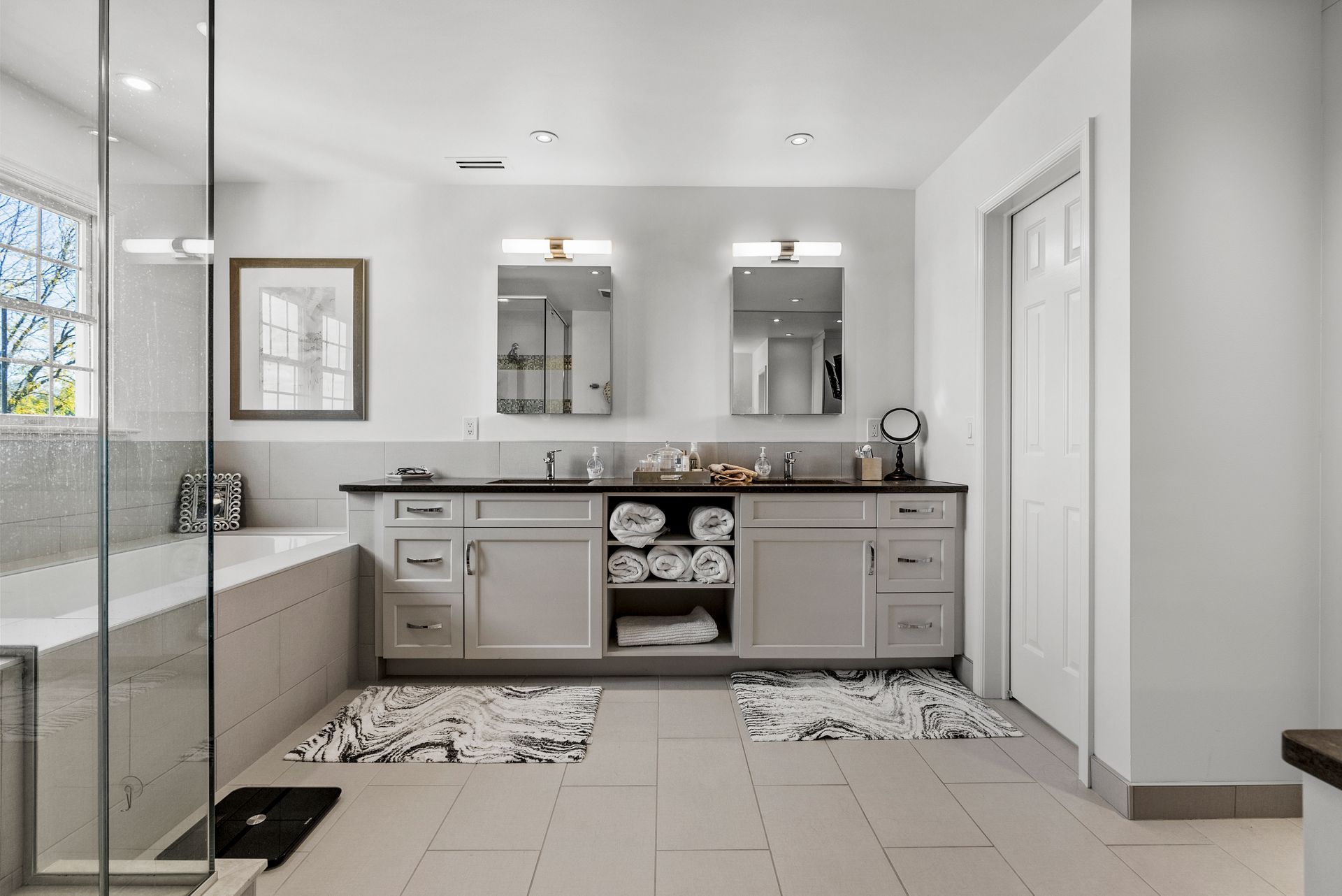 Spacious, bright bathroom with double vanity, bathtub, and large glass shower. Neutral tones and decorative rugs.