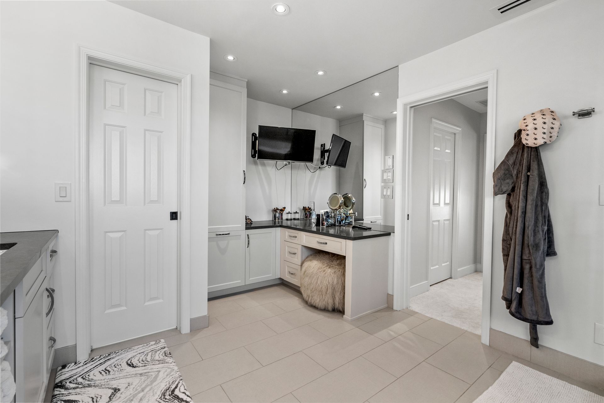 Bathroom with vanity, TV, and coat rack, featuring white walls, light-colored tile, and a fluffy stool.