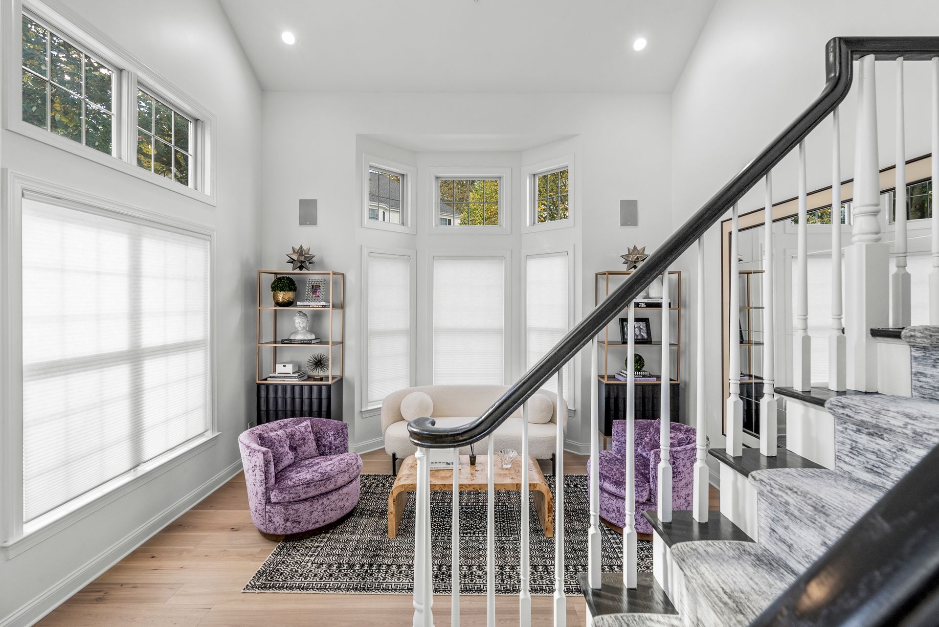 Bright white living room with stairs. Light wood floors, purple chairs, white sofa, and a bay window.