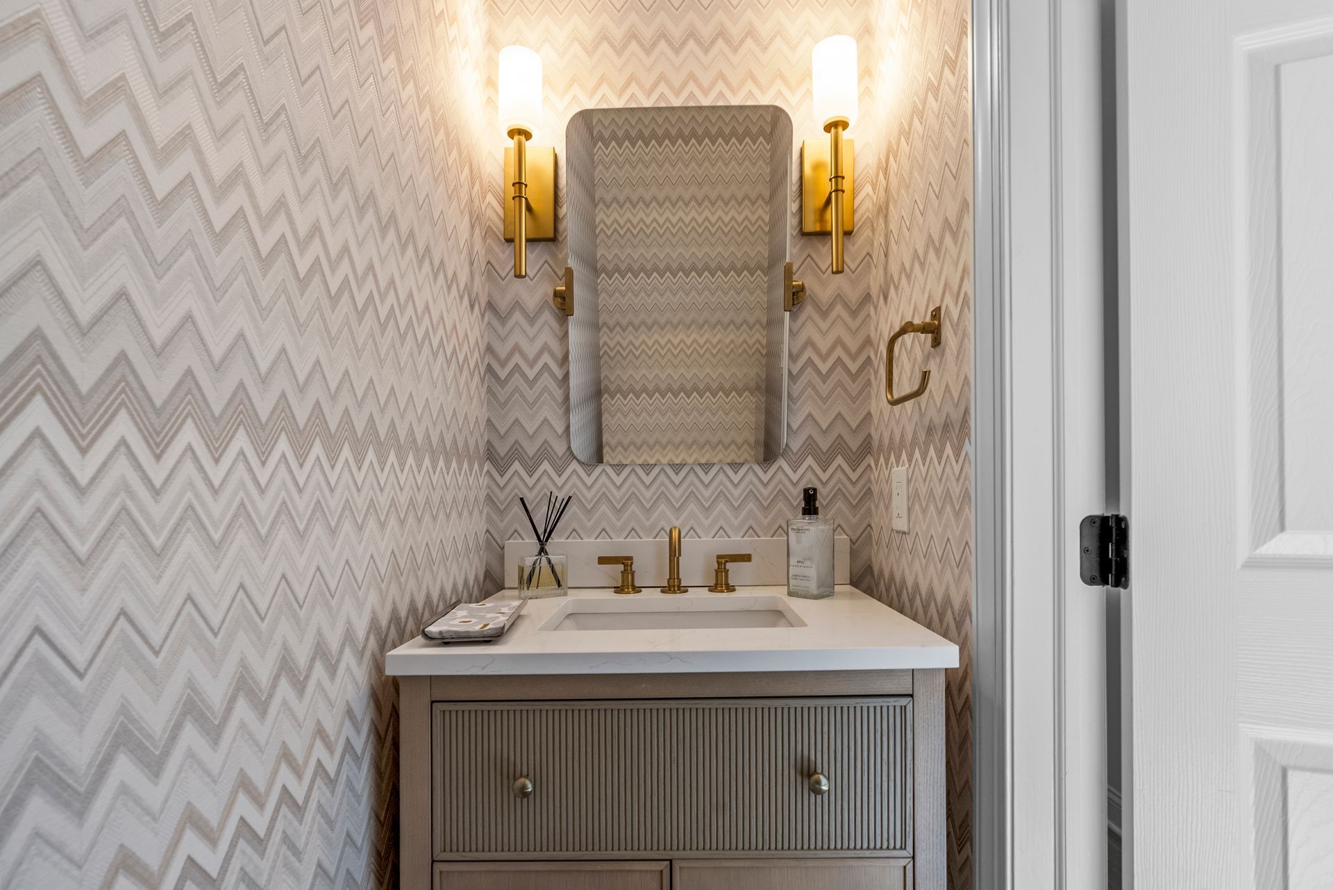 Bathroom with patterned wallpaper, gold fixtures, a light-colored vanity, and a mirror.