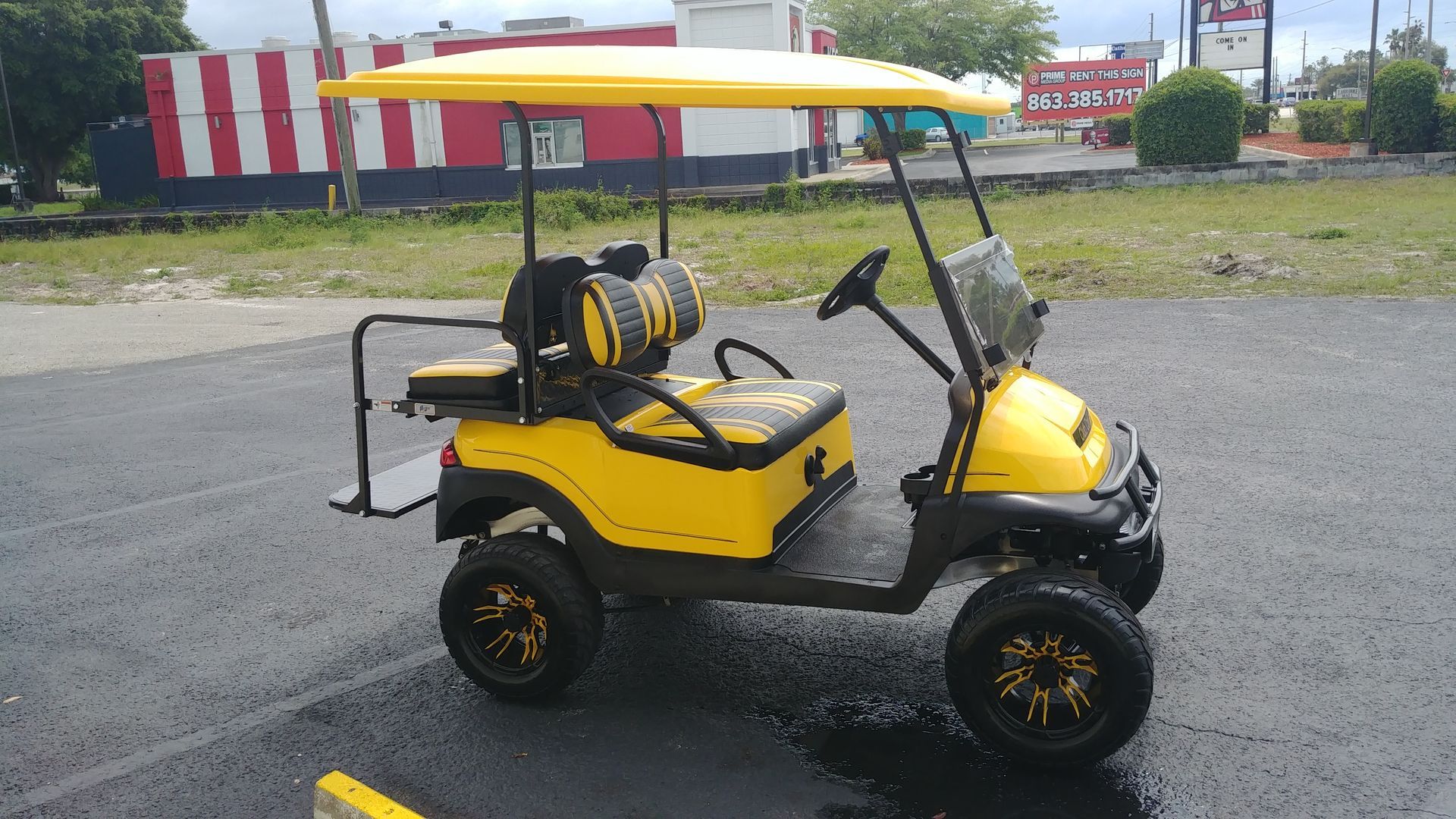 A yellow golf cart with black accents and custom wheels parked on an asphalt lot in front of a building.