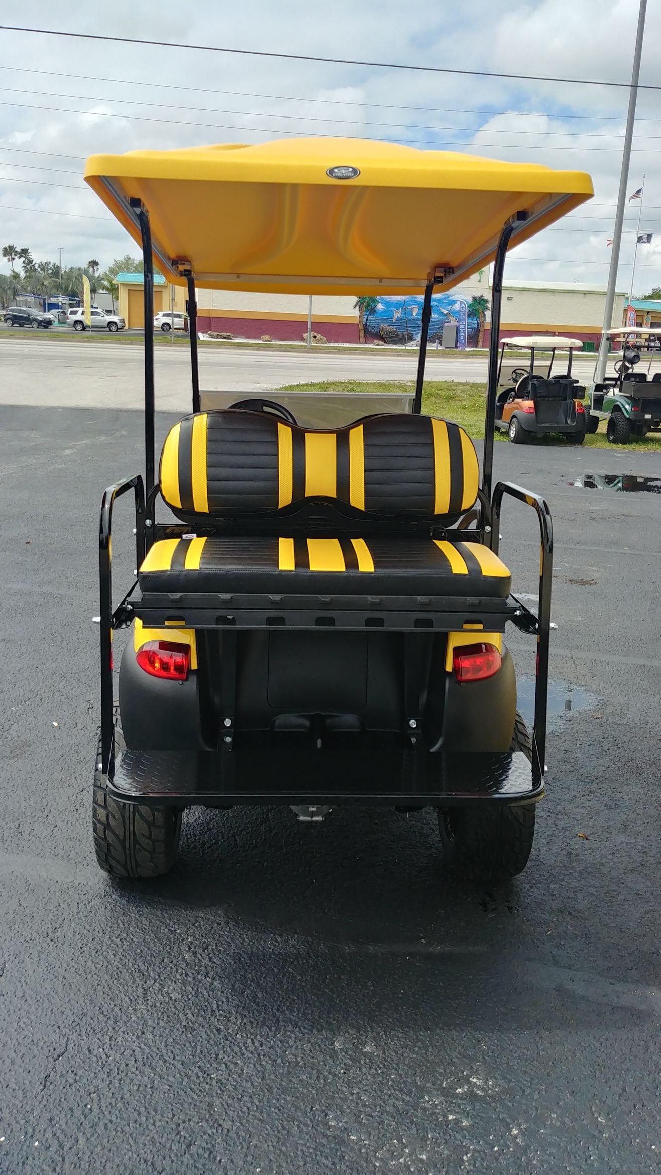 Rear view of a yellow and black golf cart parked on an asphalt lot under a cloudy sky.