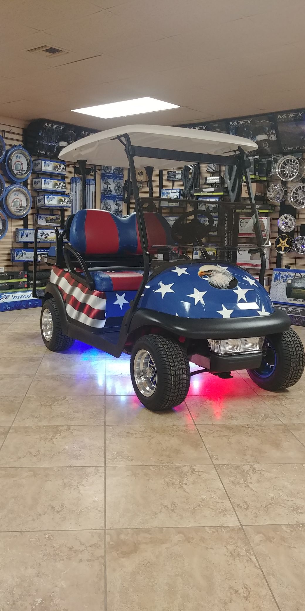 A golf cart with a patriotic American flag and star paint job parked indoors with glowing blue and red under-glow lights.
