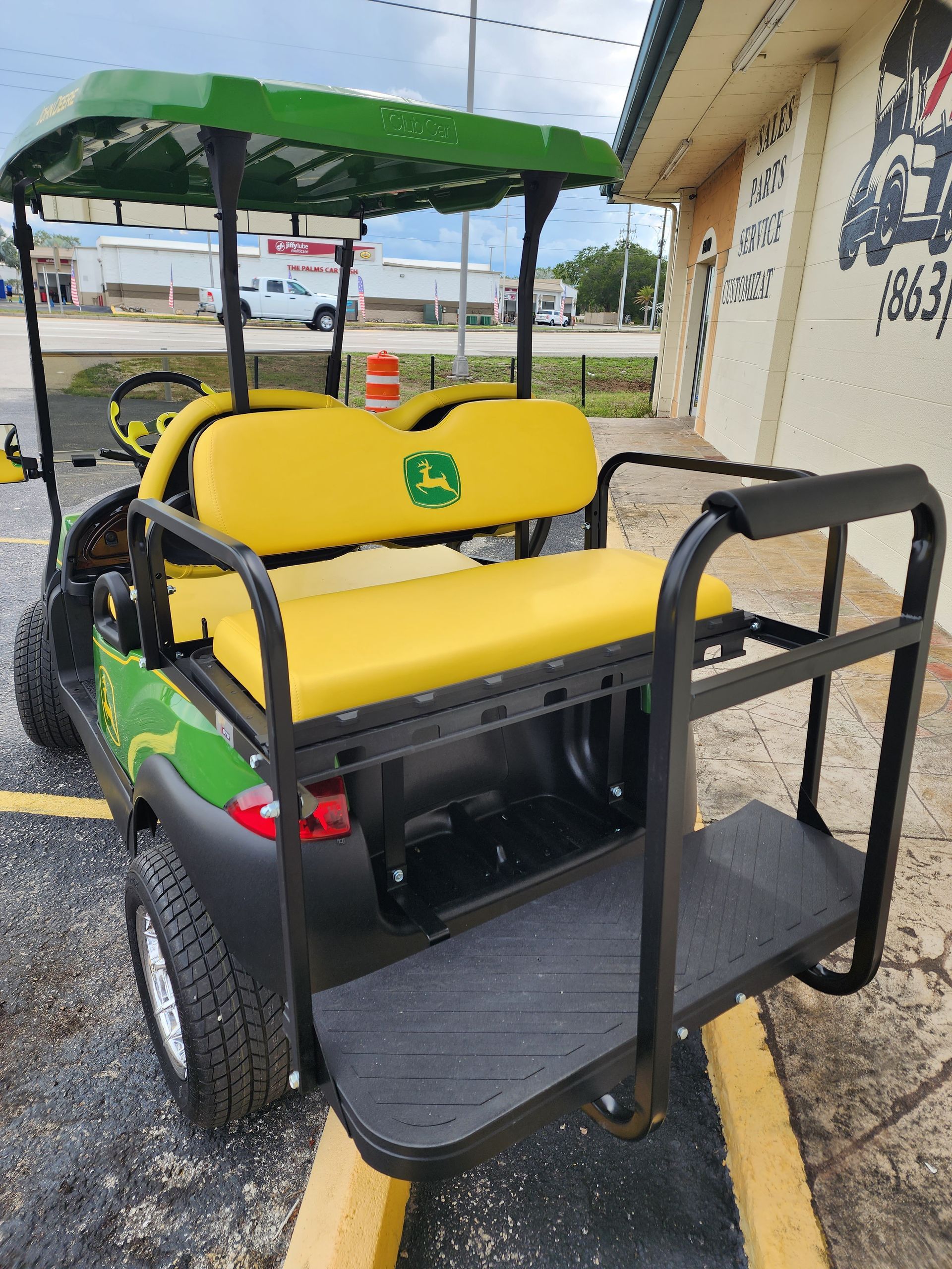 A John Deere-branded golf cart with a yellow rear seat and a black flatbed, parked outside a building.