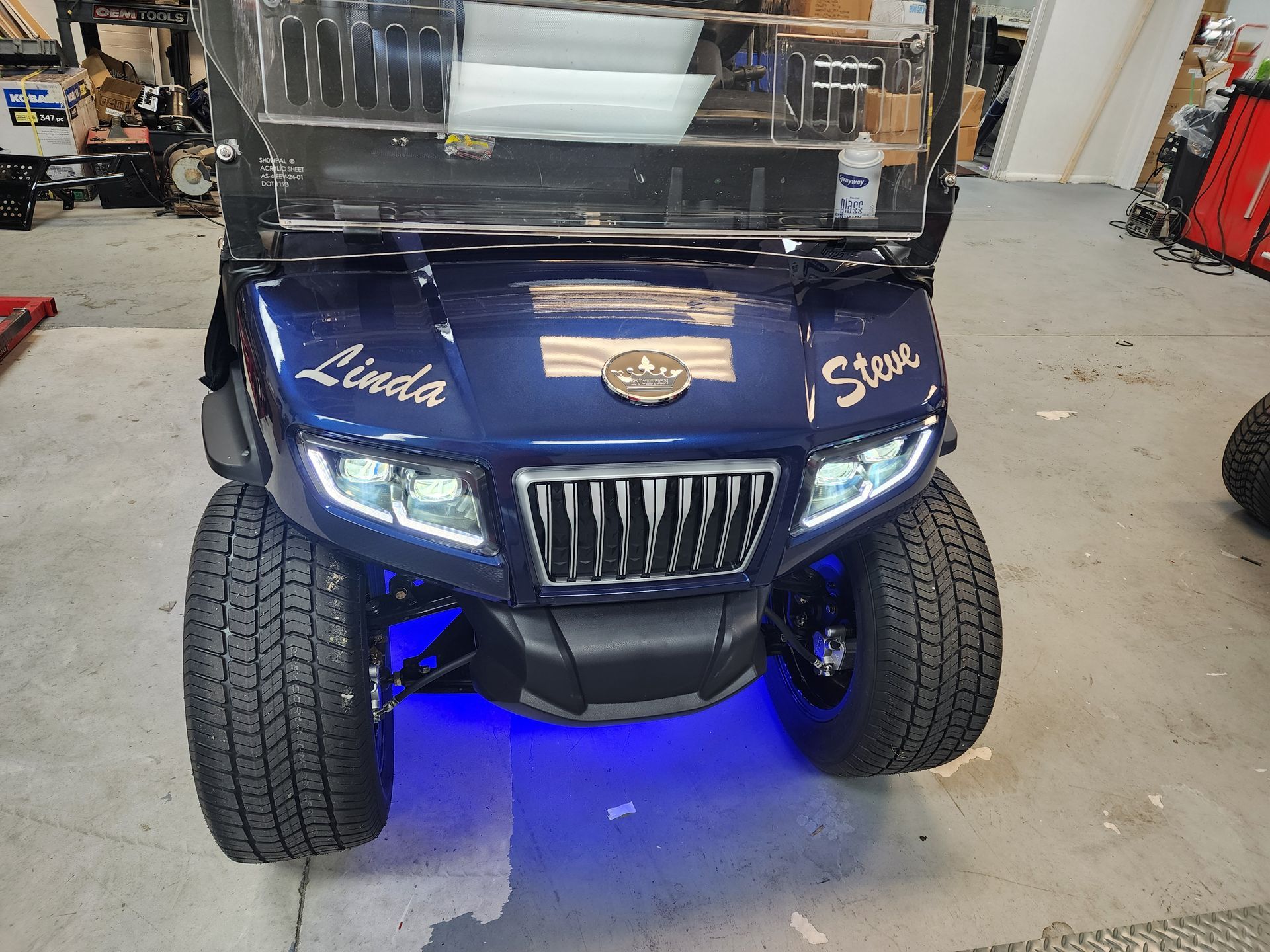 A dark blue golf cart with LED headlights and blue underglow, parked inside a shop.