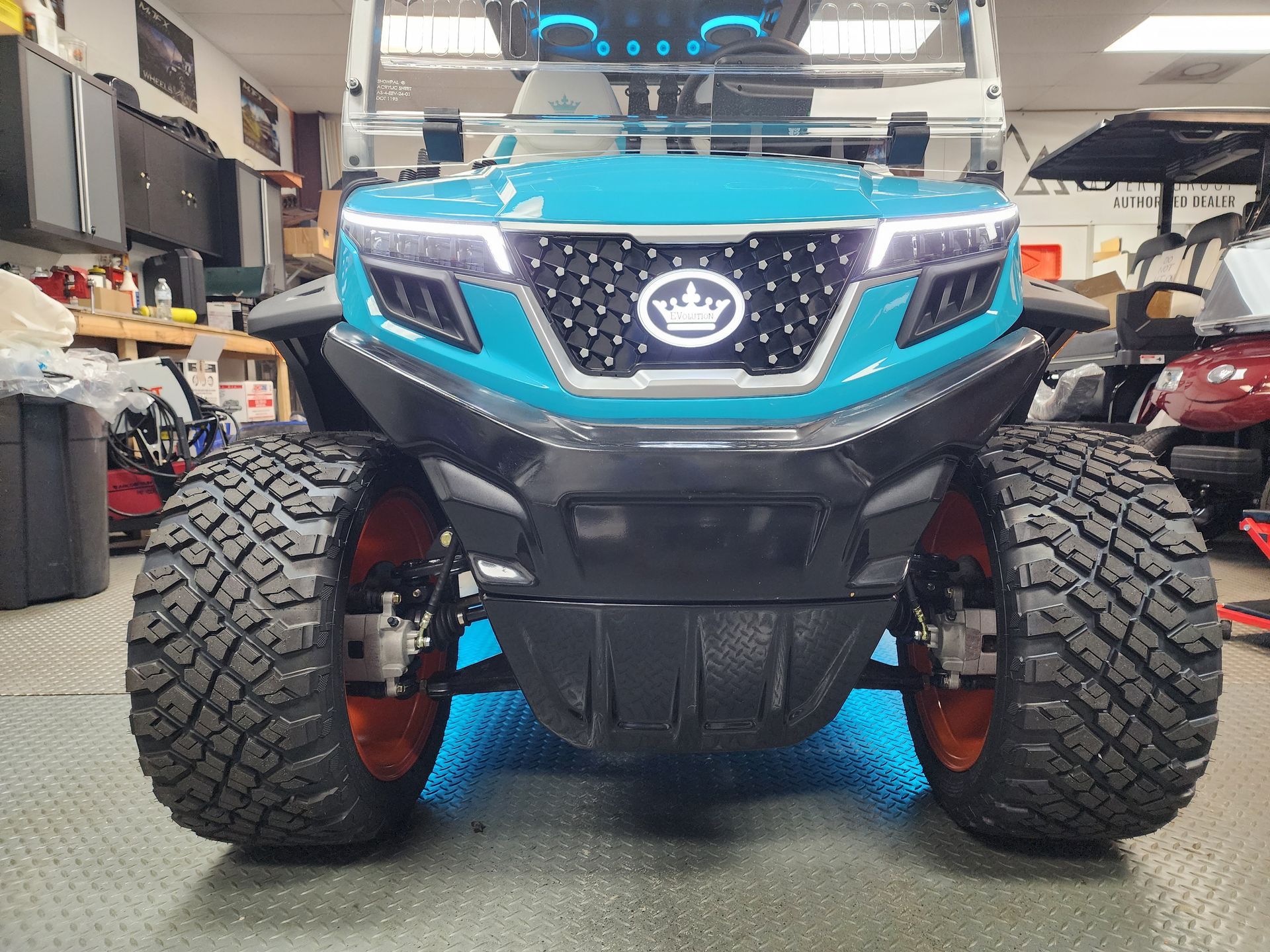 Front view of a bright teal golf cart with a prominent crown logo, black grille, and chunky tires in a showroom.