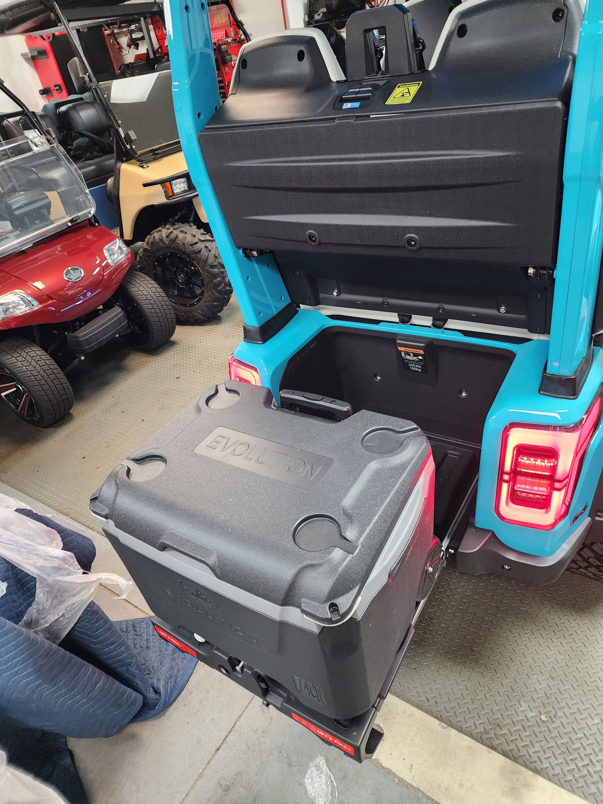 A teal utility vehicle in a shop, with a black cooler strapped to a rear rack and a red UTV parked in the background.