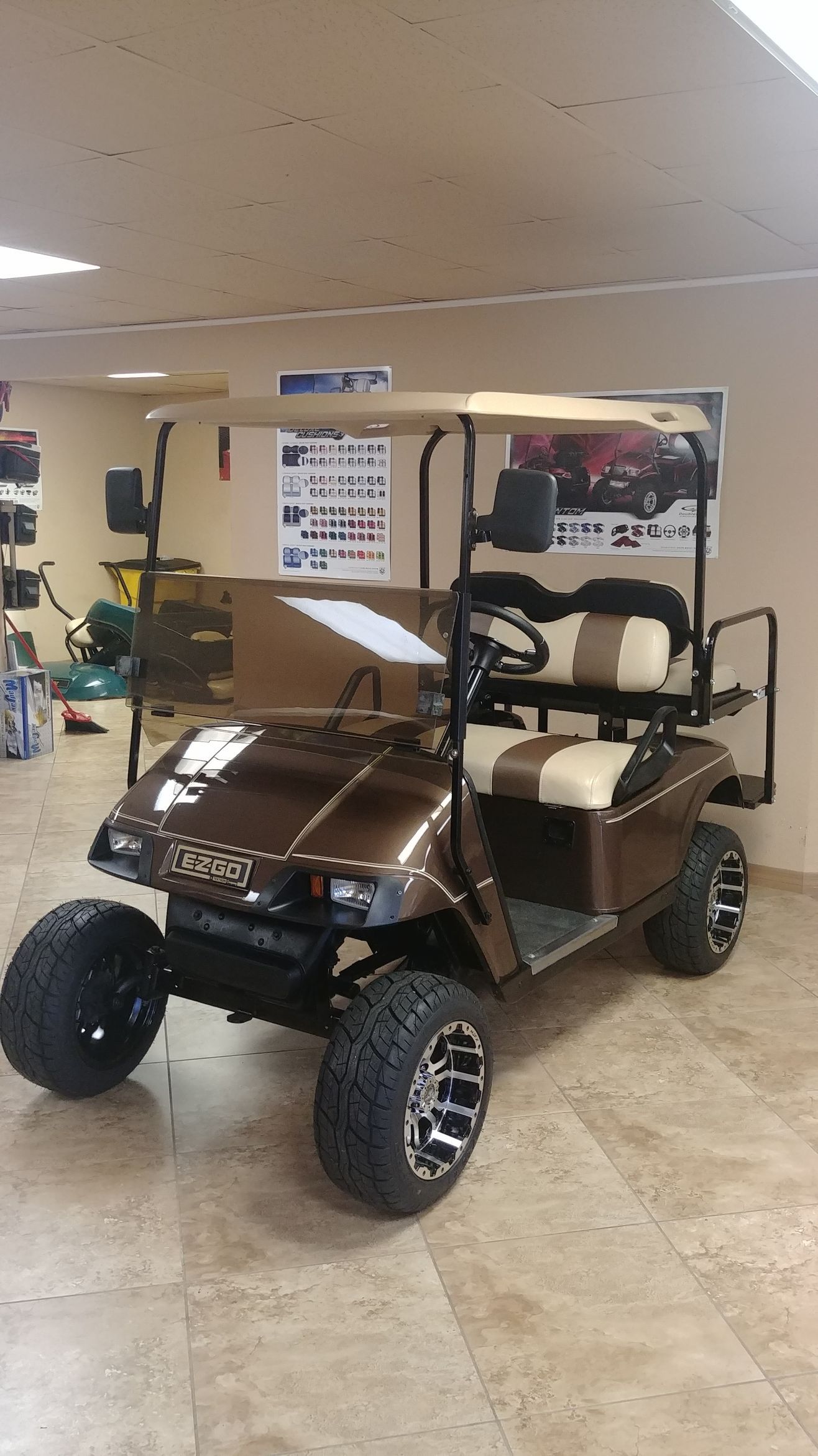 Brown golf cart with off-road tires, tan seats, and a windshield indoors.