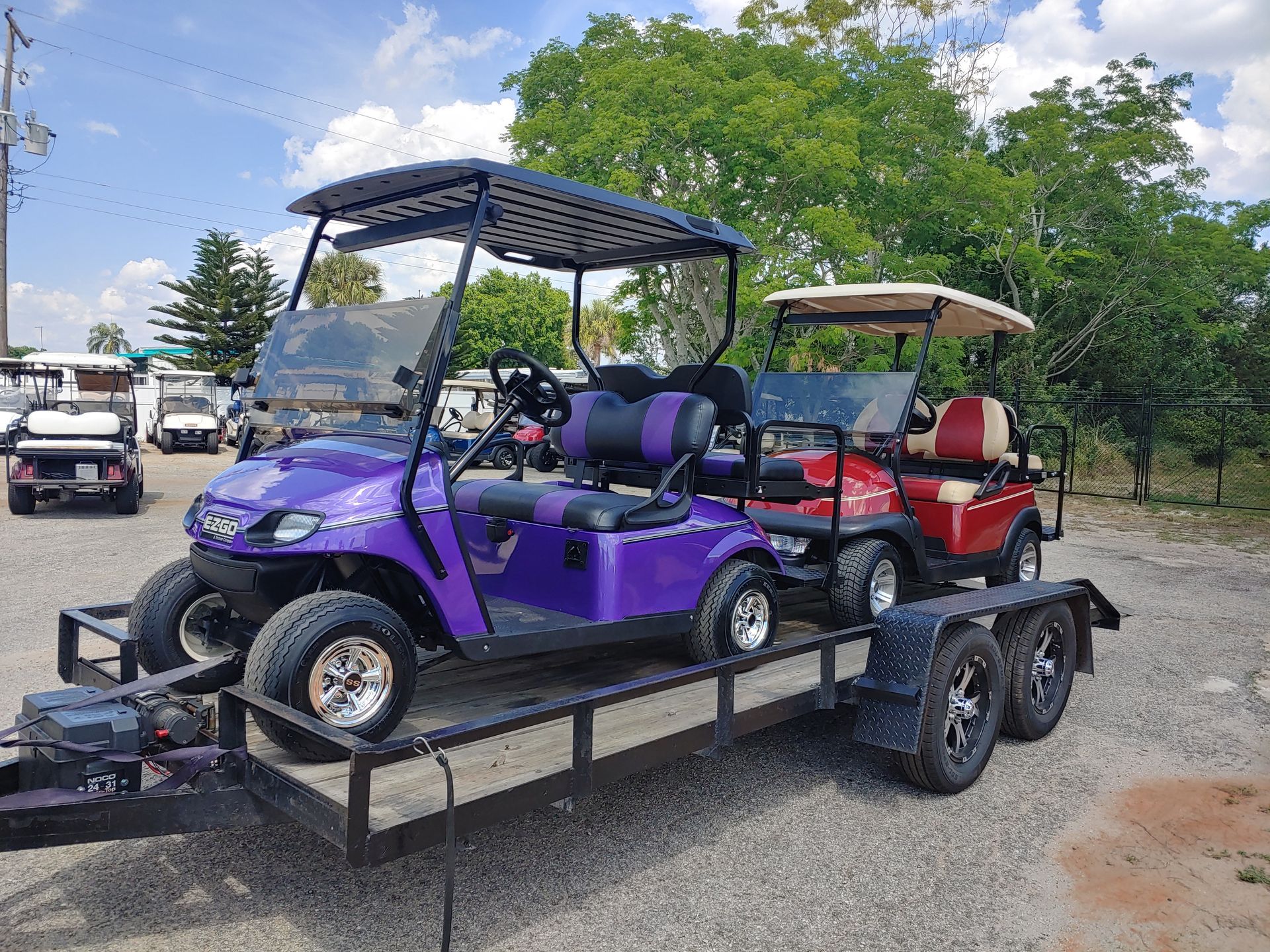Three golf carts on a black trailer, outdoors. Purple, red, and white carts; sunny day.