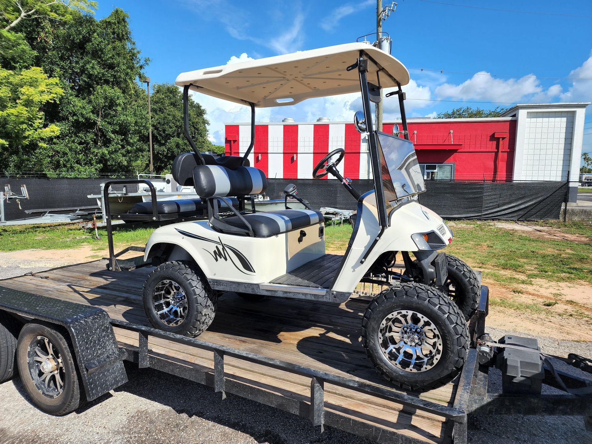 Cream-colored golf cart with lift kit and black accents on a trailer in front of a red and white building.