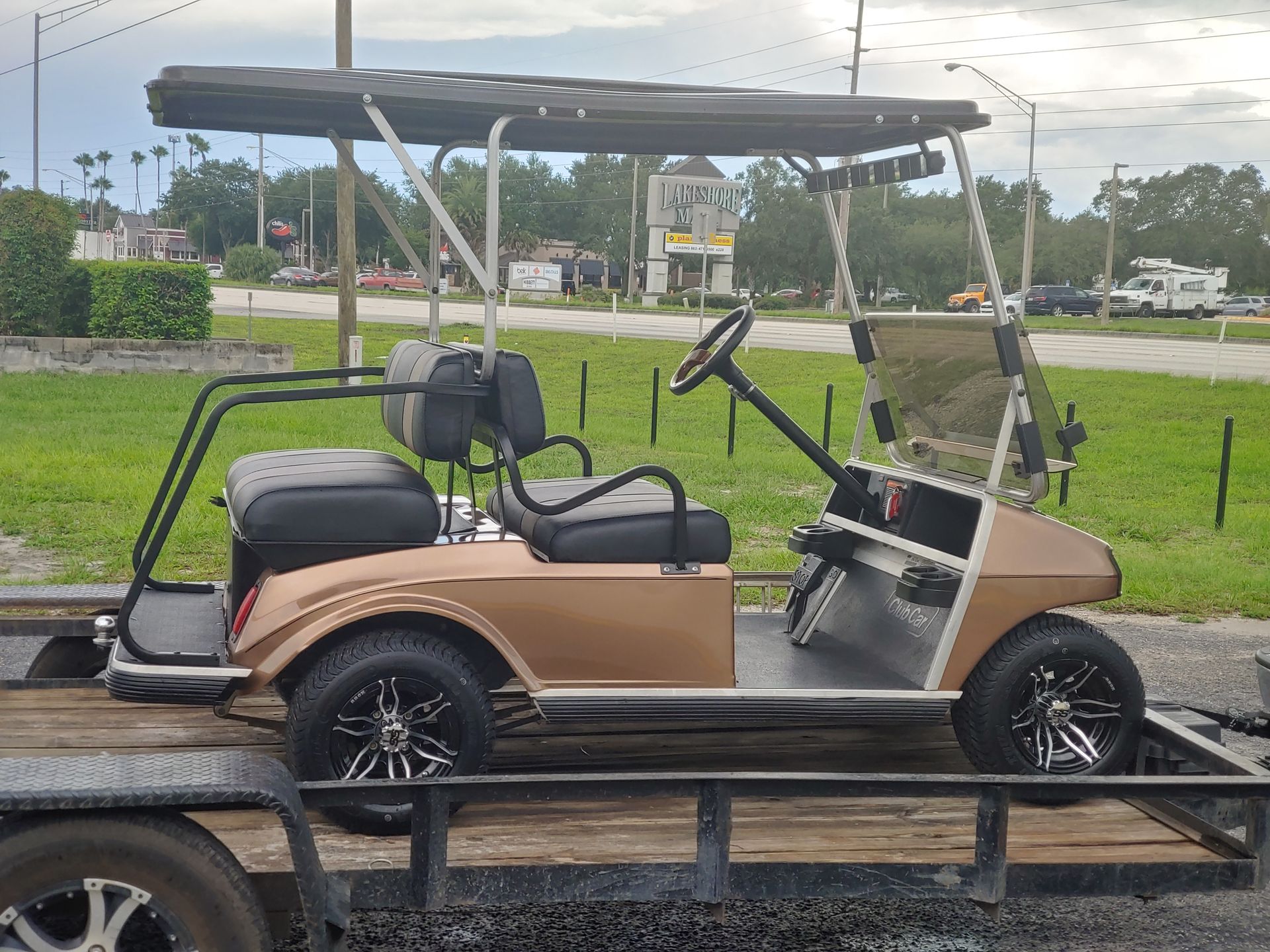 Tan golf cart on a trailer, with black wheels, seats, and canopy, parked on a road.