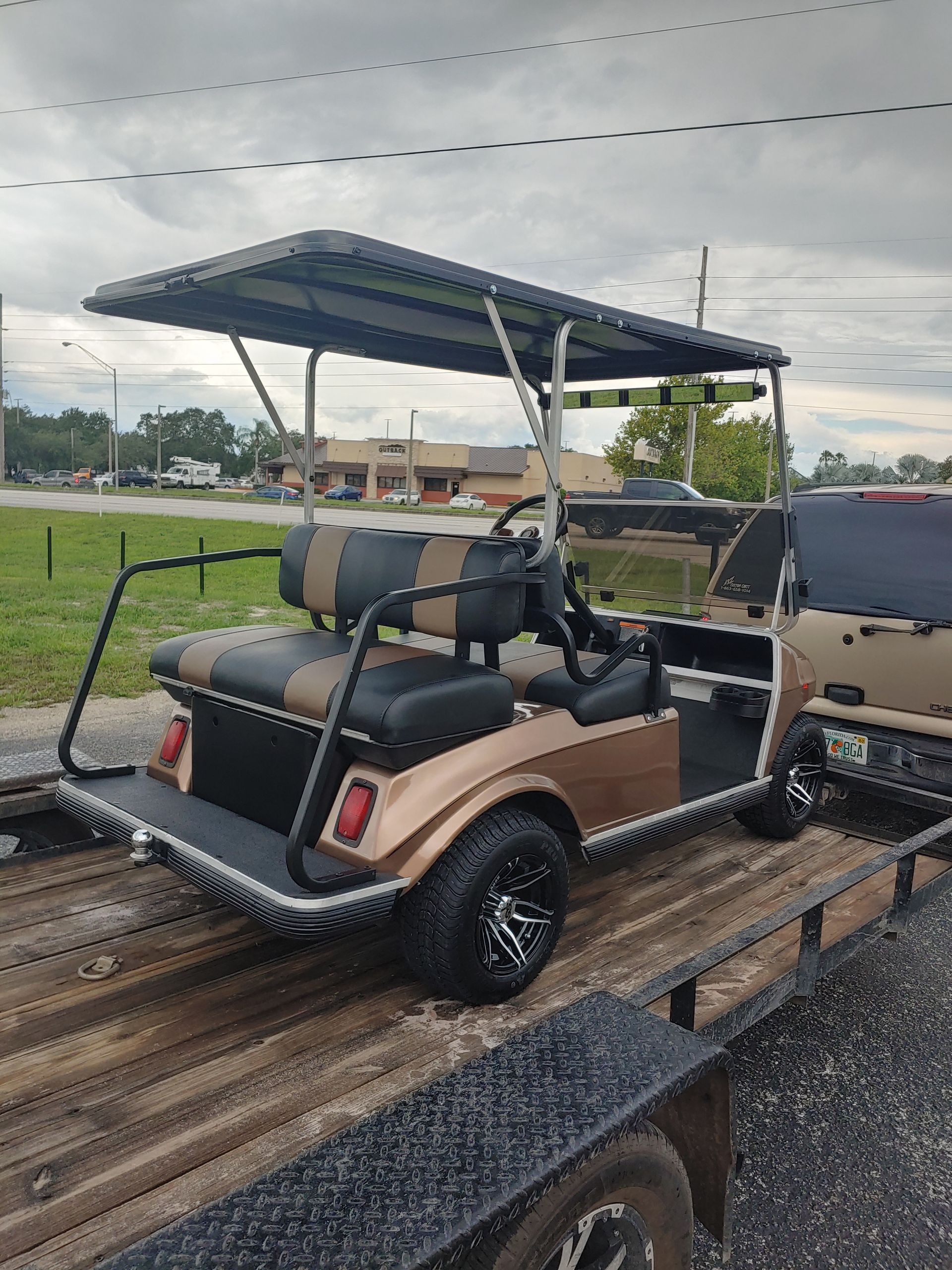 Gold and black golf cart on a trailer, black wheels, canopy, and two-tone seats.