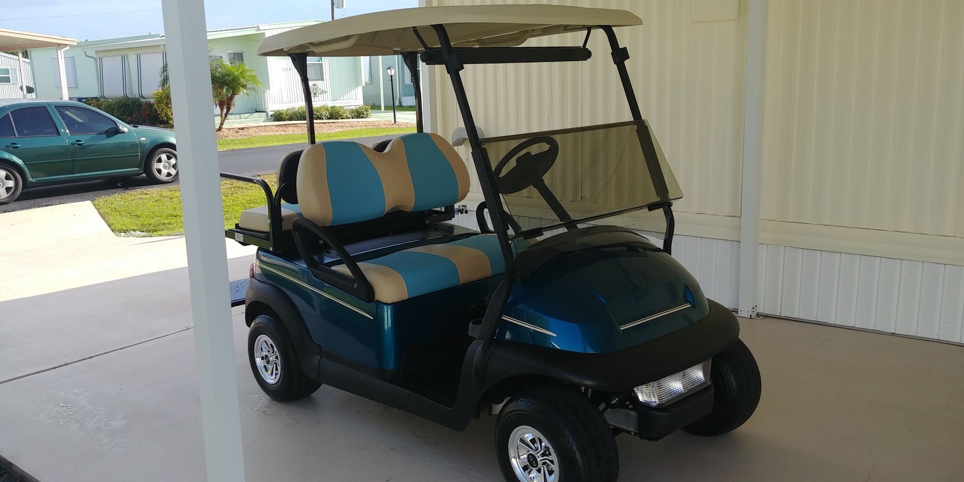 A blue golf cart with tan and blue seats parked under a beige awning. A green car is in the background.