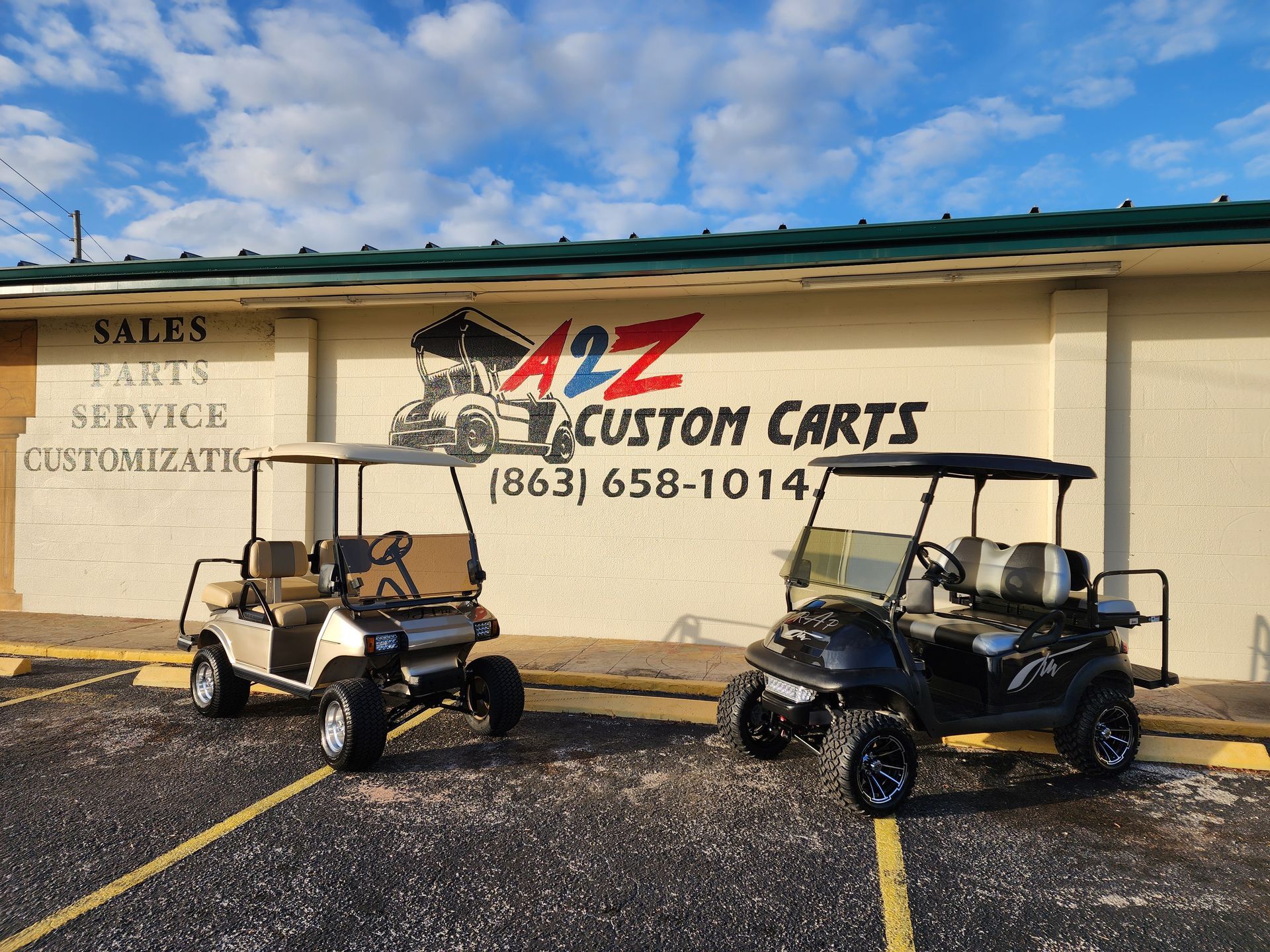 Two golf carts parked in front of A2Z Custom Carts storefront. Building is tan and green, with blue sky.