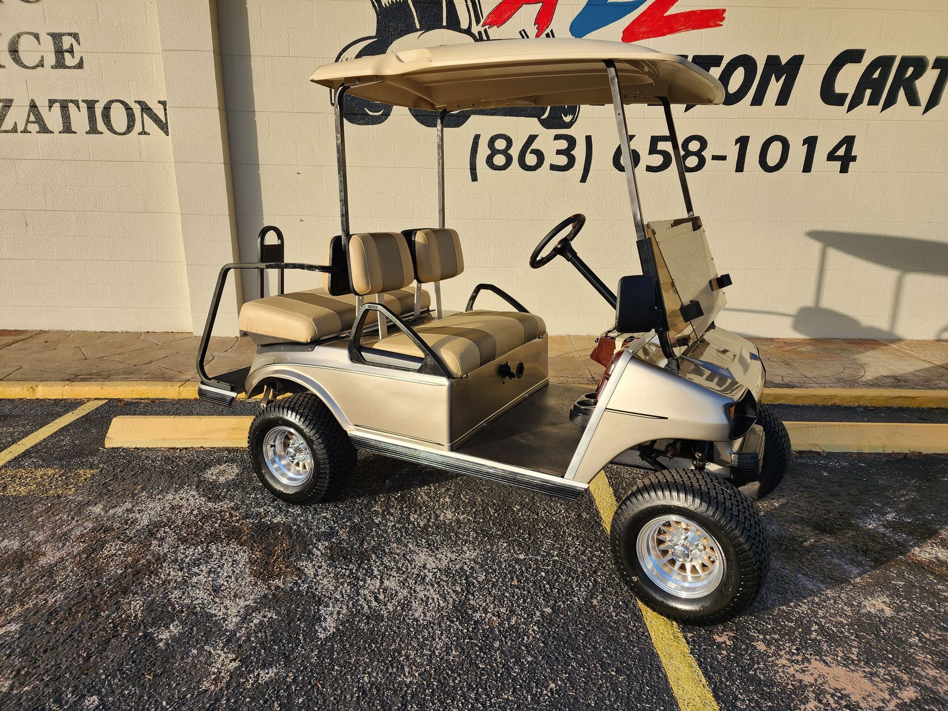 Tan golf cart parked outside, shiny wheels, beige canopy, phone number visible.