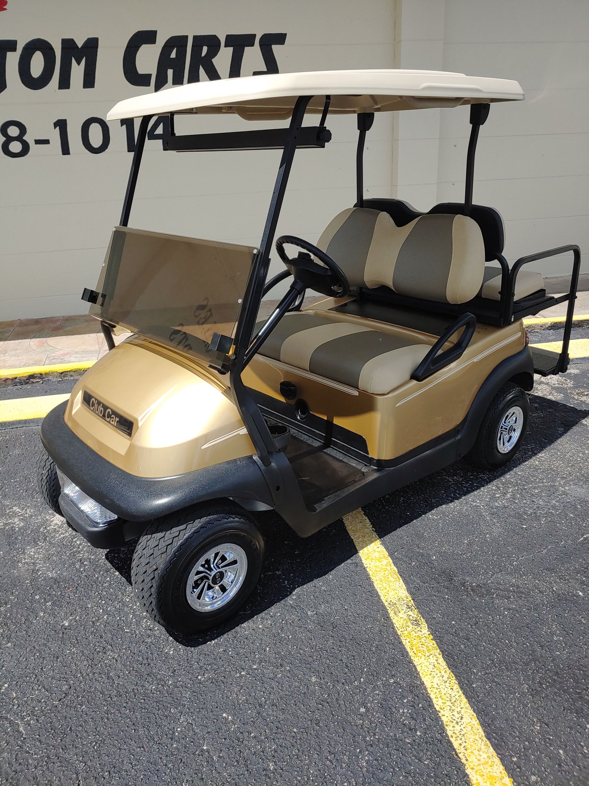 Gold golf cart with tan roof, seats, and windshield. Parked on asphalt.