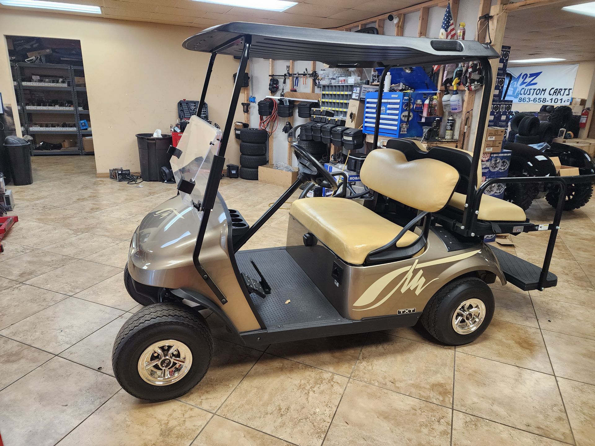 Tan golf cart with black roof, tan seats, and silver wheels inside a shop.