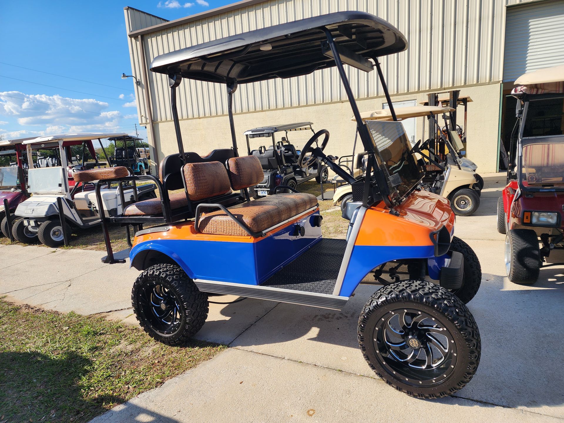 Orange and blue golf cart with large wheels parked outside a building, other carts in background.