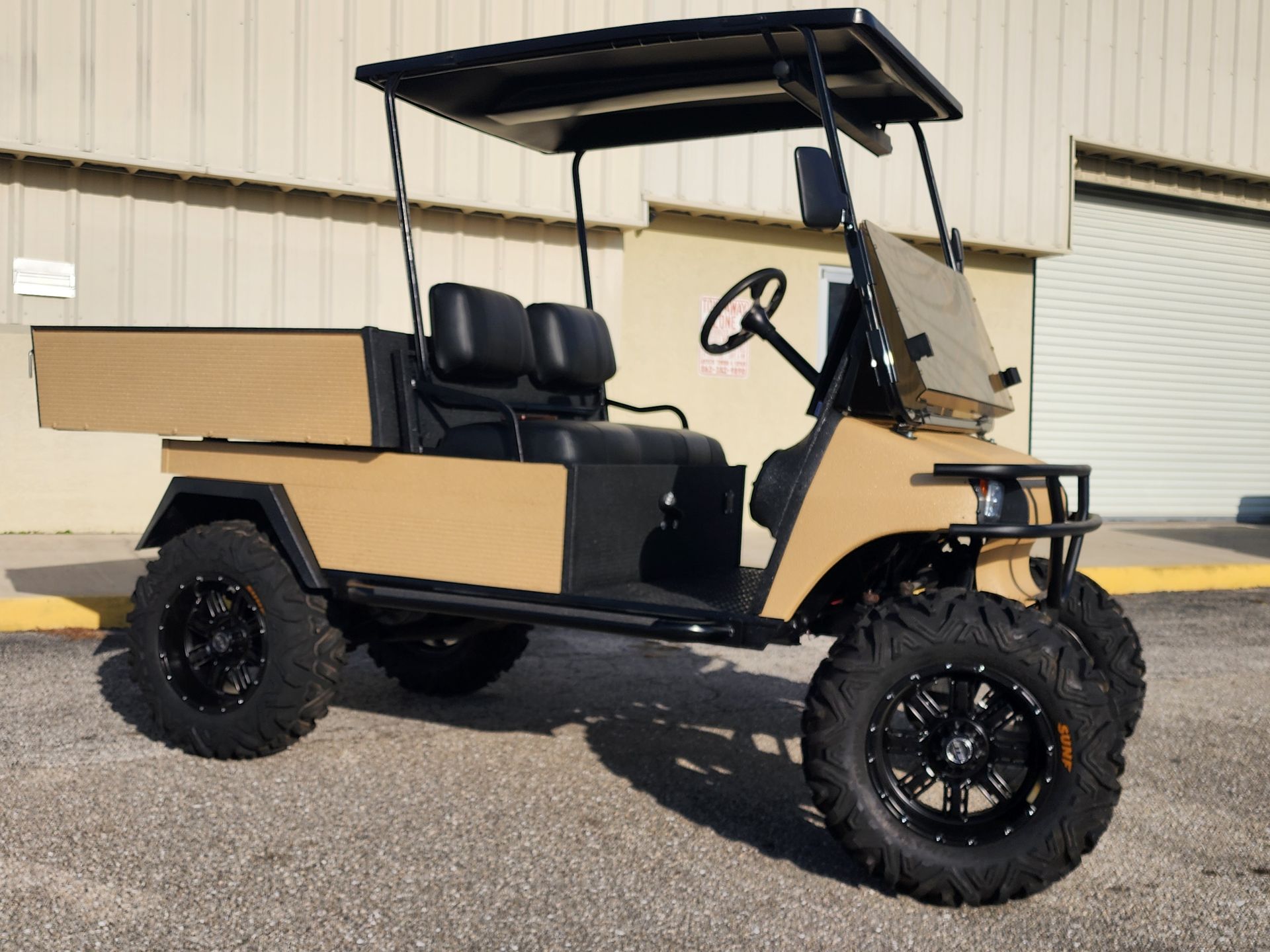 Tan custom golf cart with black wheels, seats, and roof. It has a cargo bed and a windshield.