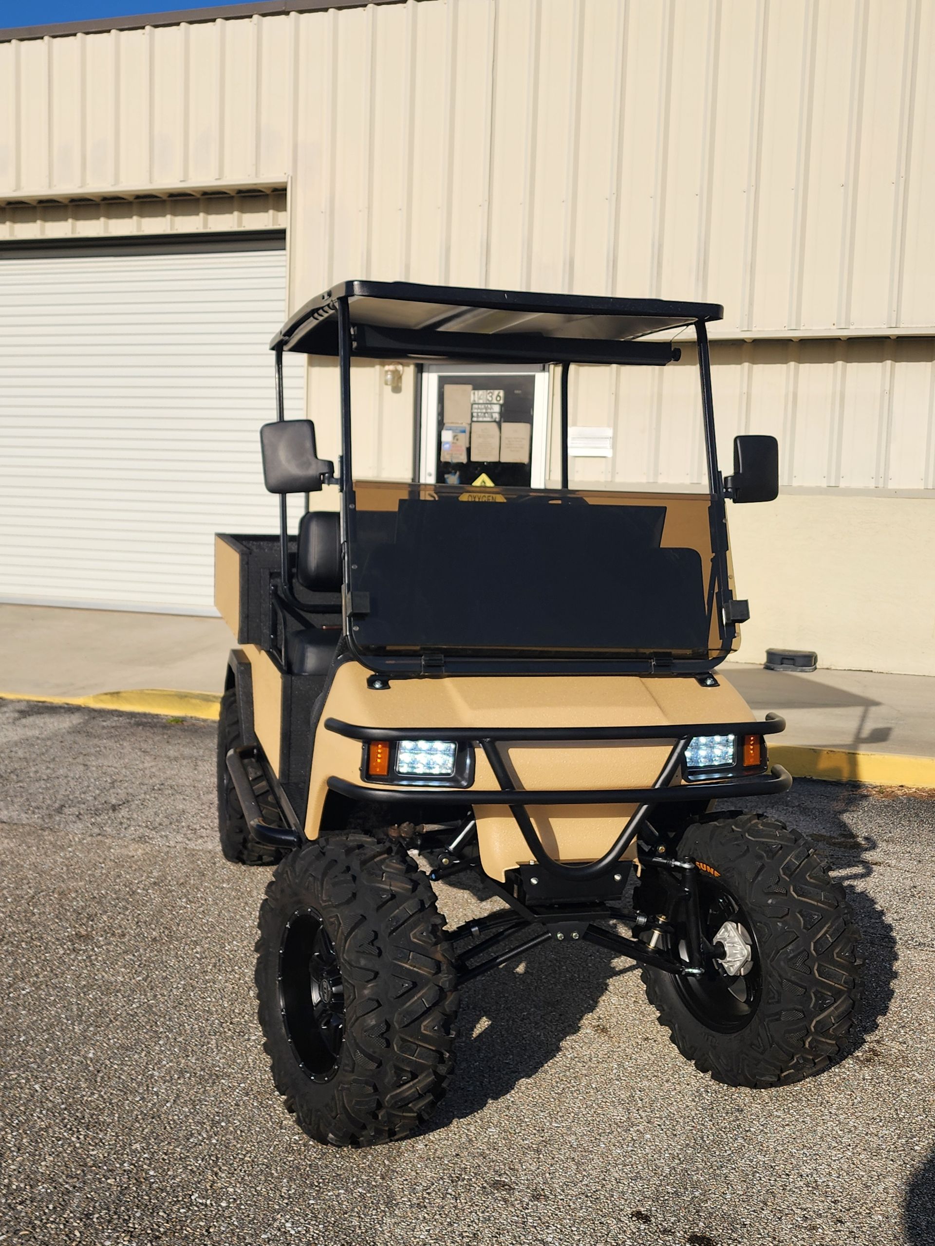 Tan off-road golf cart with black accents, large tires, and a protective front bumper, parked on gravel.