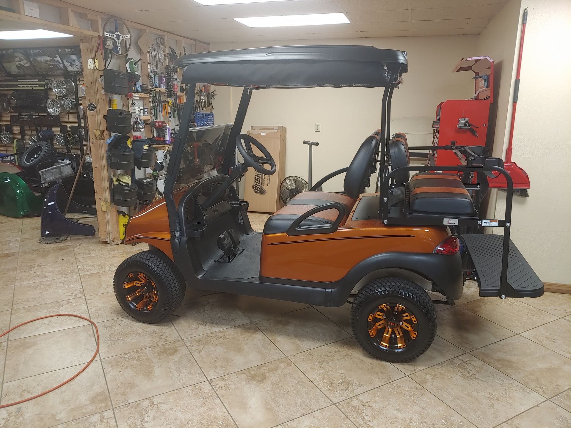 Orange golf cart with black roof, custom wheels, and rear seat, parked indoors.