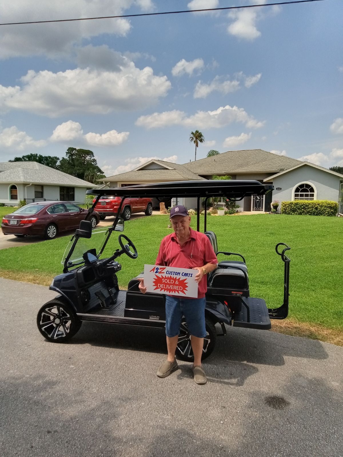 Man with sign stands in front of a black golf cart on a residential street under a blue sky.