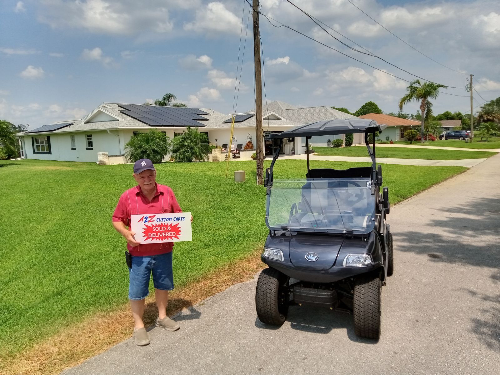Man holding a box next to a golf cart on a residential street with houses and solar panels. Blue sky.