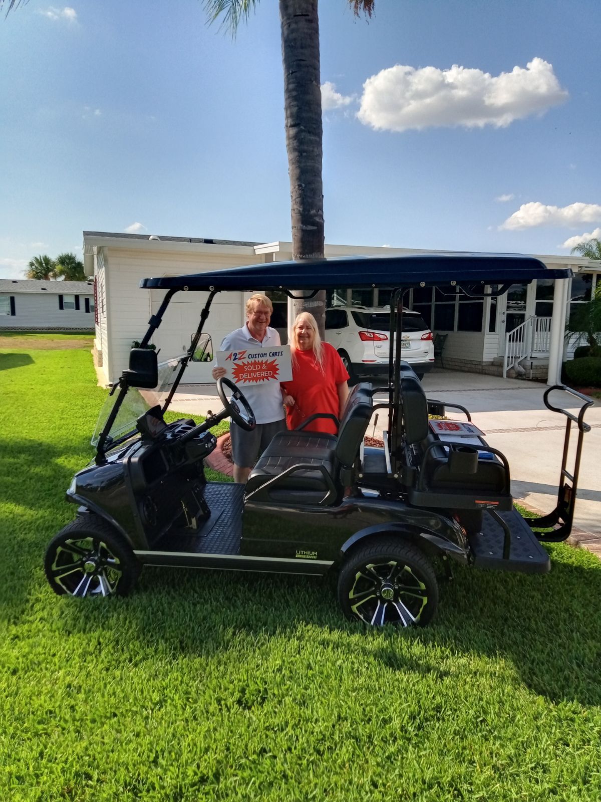 A couple stands next to a new black golf cart on a lawn; bright sun.