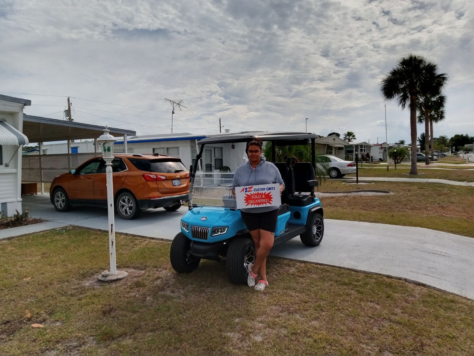 Woman holding box, standing beside blue golf cart. Houses and orange car in background, cloudy sky.