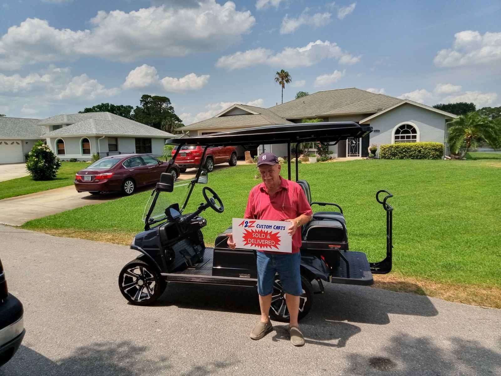 Man with golf cart, holding a sign. Sunny day, houses and red car in background.