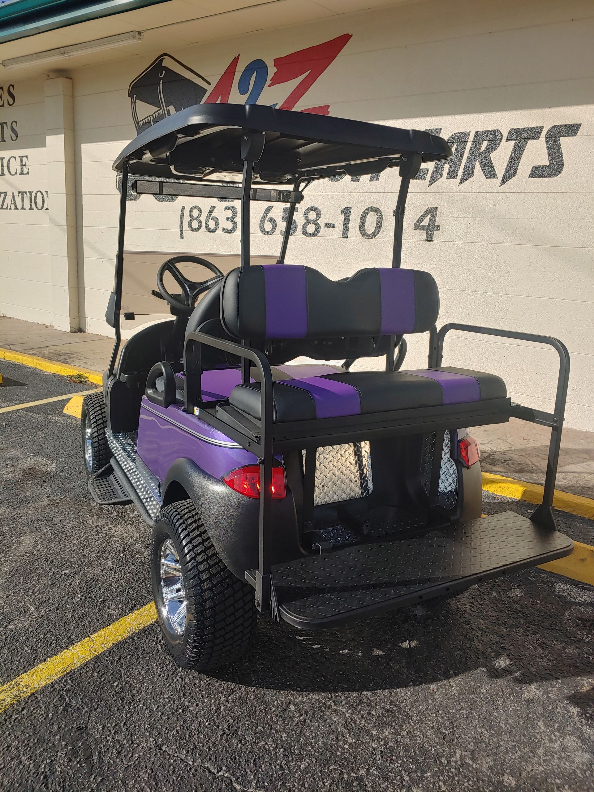 Purple and black golf cart with a rear seat and utility platform, parked outdoors.