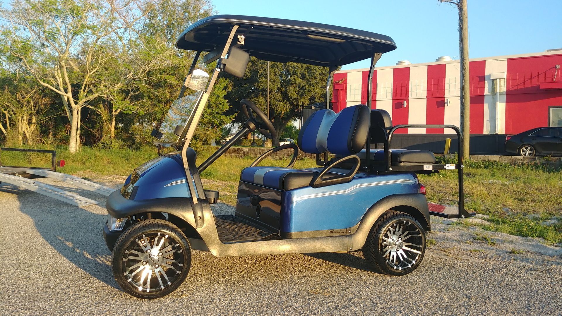 Blue golf cart with custom wheels, parked on gravel. In the background is greenery and a red and white building.