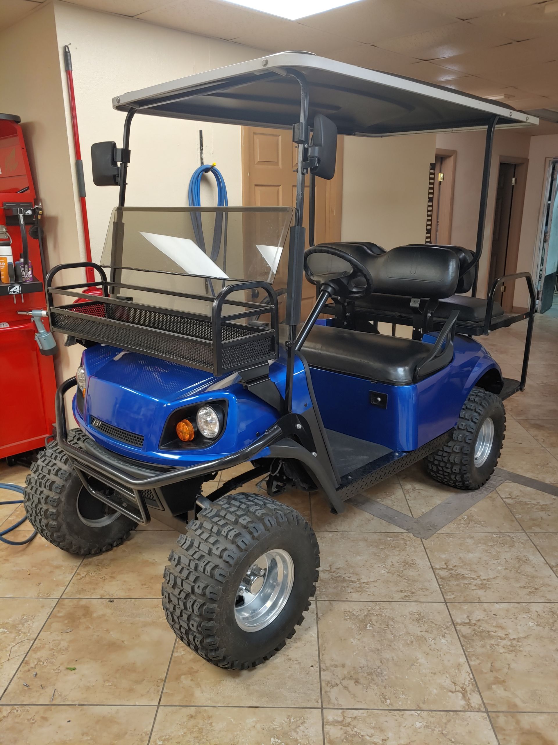 Blue golf cart with off-road tires, a canopy, and a front basket parked inside a building.
