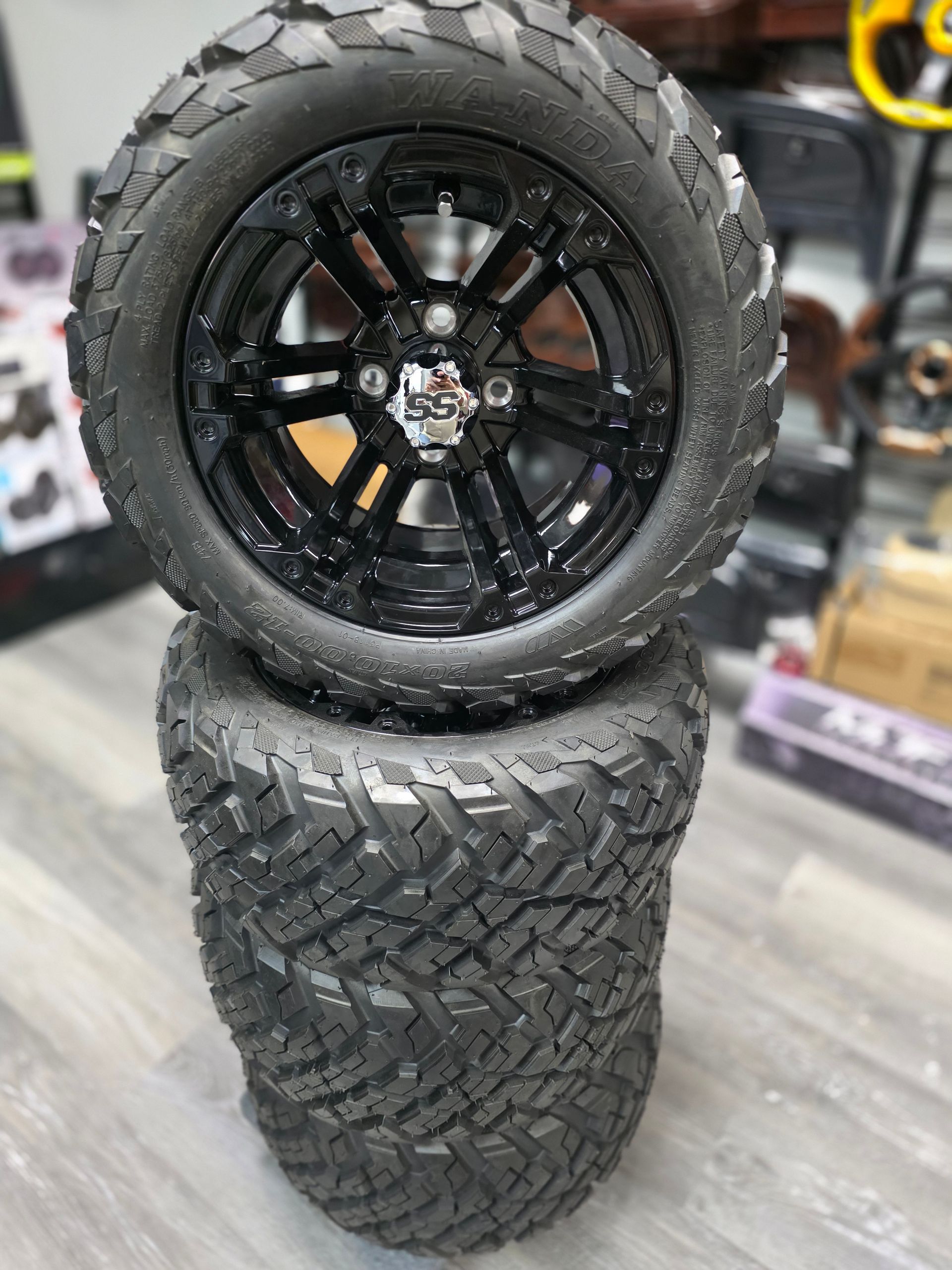 Two black off-road golf cart tires with glossy black rims stacked vertically on a shop floor.