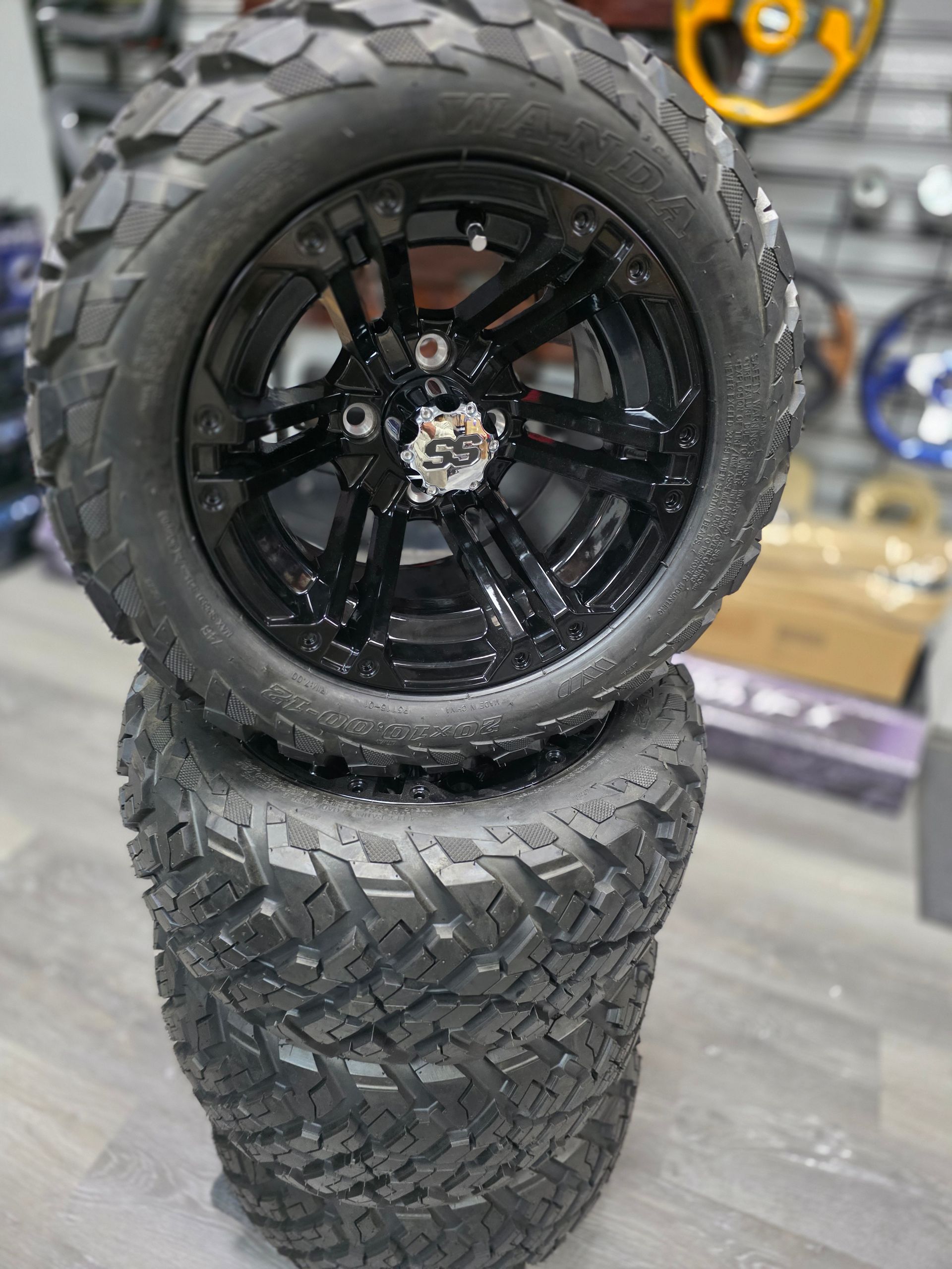 Two black, rugged off-road tires stacked on top of each other in a shop with automotive accessories in the background.