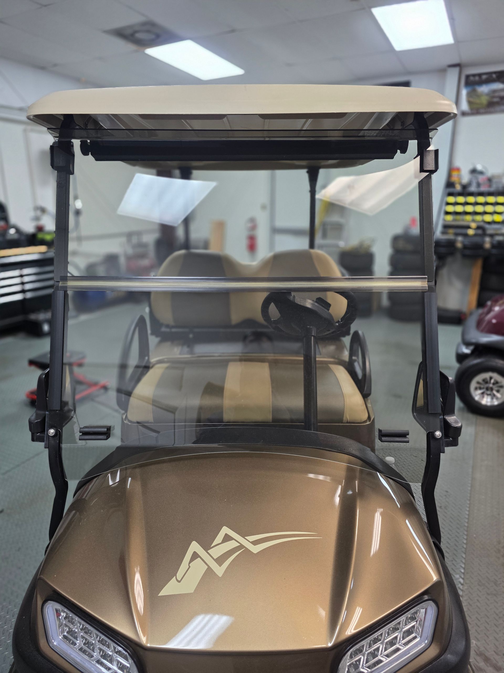 Brown golf cart indoors with clear windshield and tan roof in a workshop