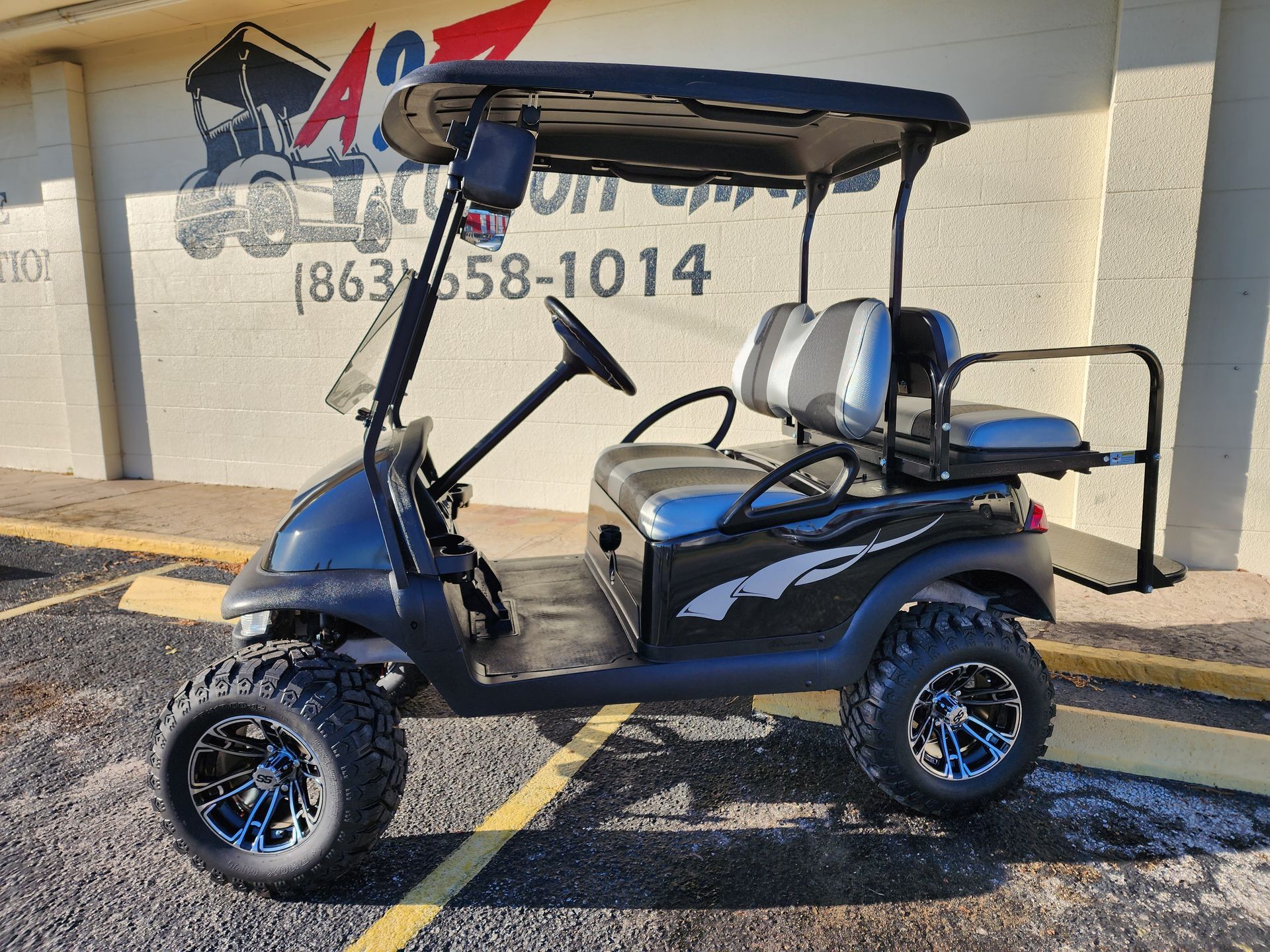 Blue golf cart with black canopy and off-road tires parked in a lot beside a wall with a painted logo.