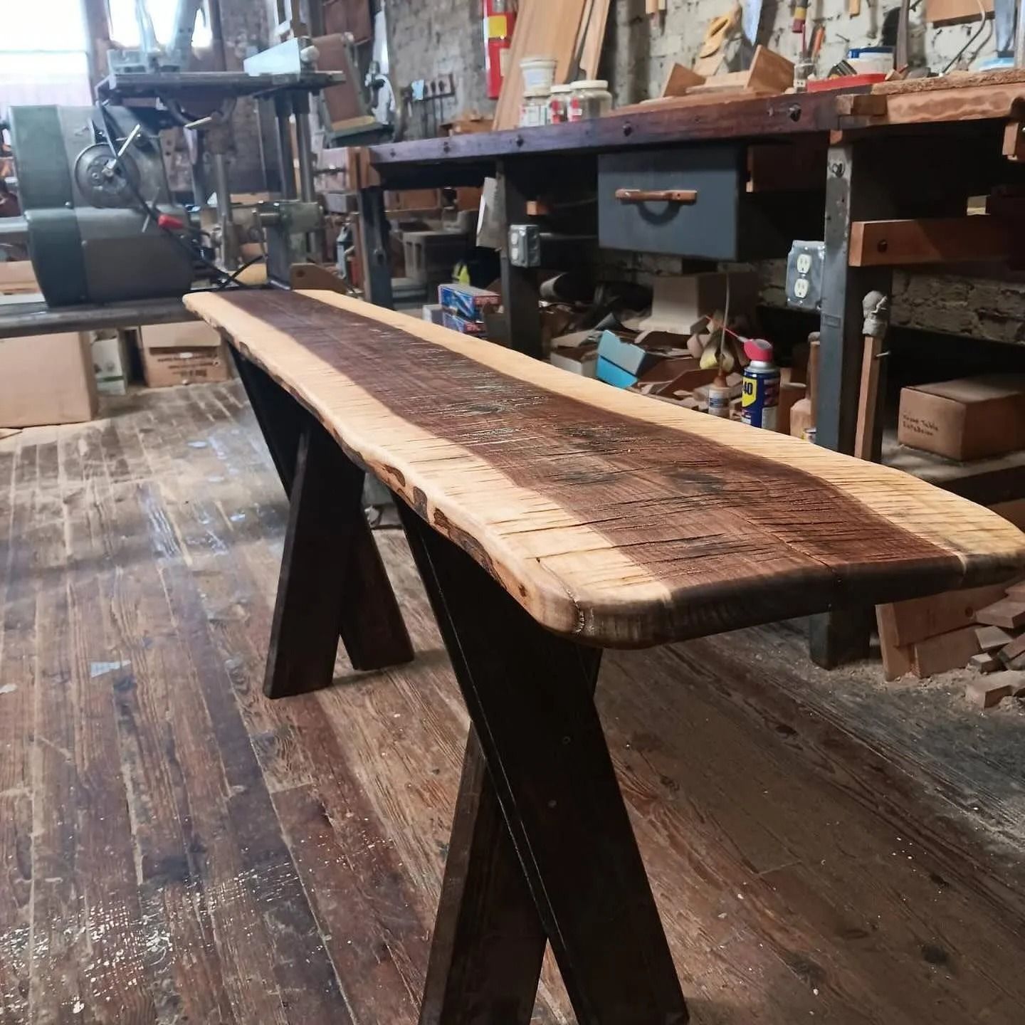 Live-edge wooden console table with dark criss-cross legs in a workshop setting.