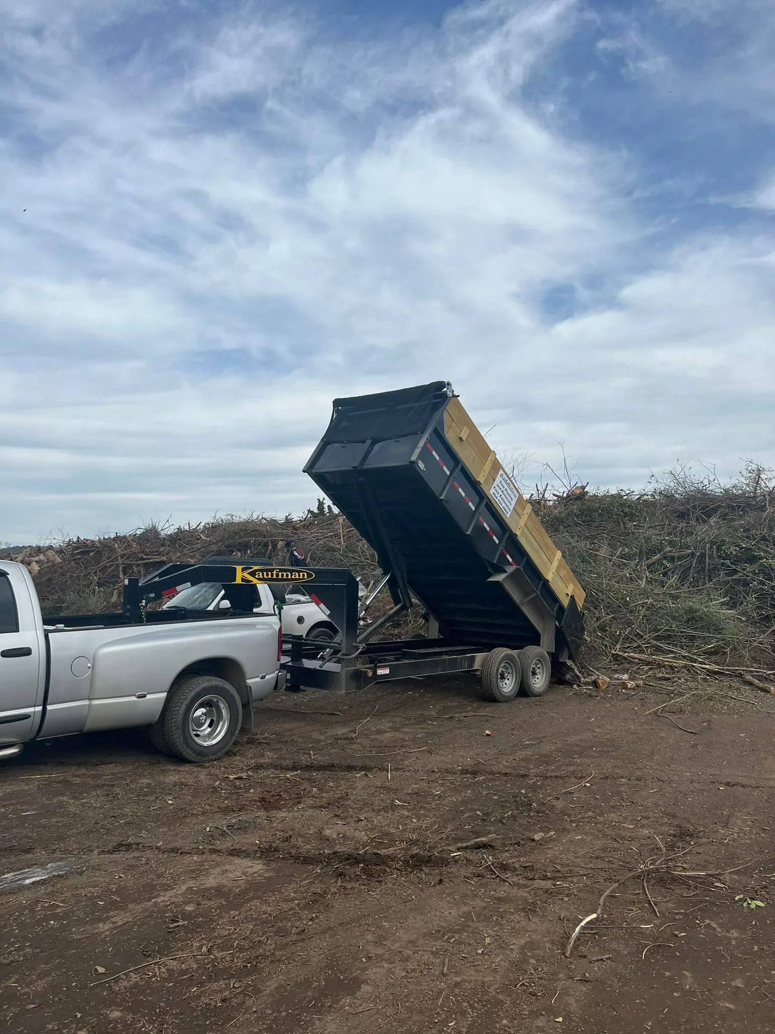 A silver pickup truck dumping debris from a black trailer onto a large pile of branches under a cloudy sky.