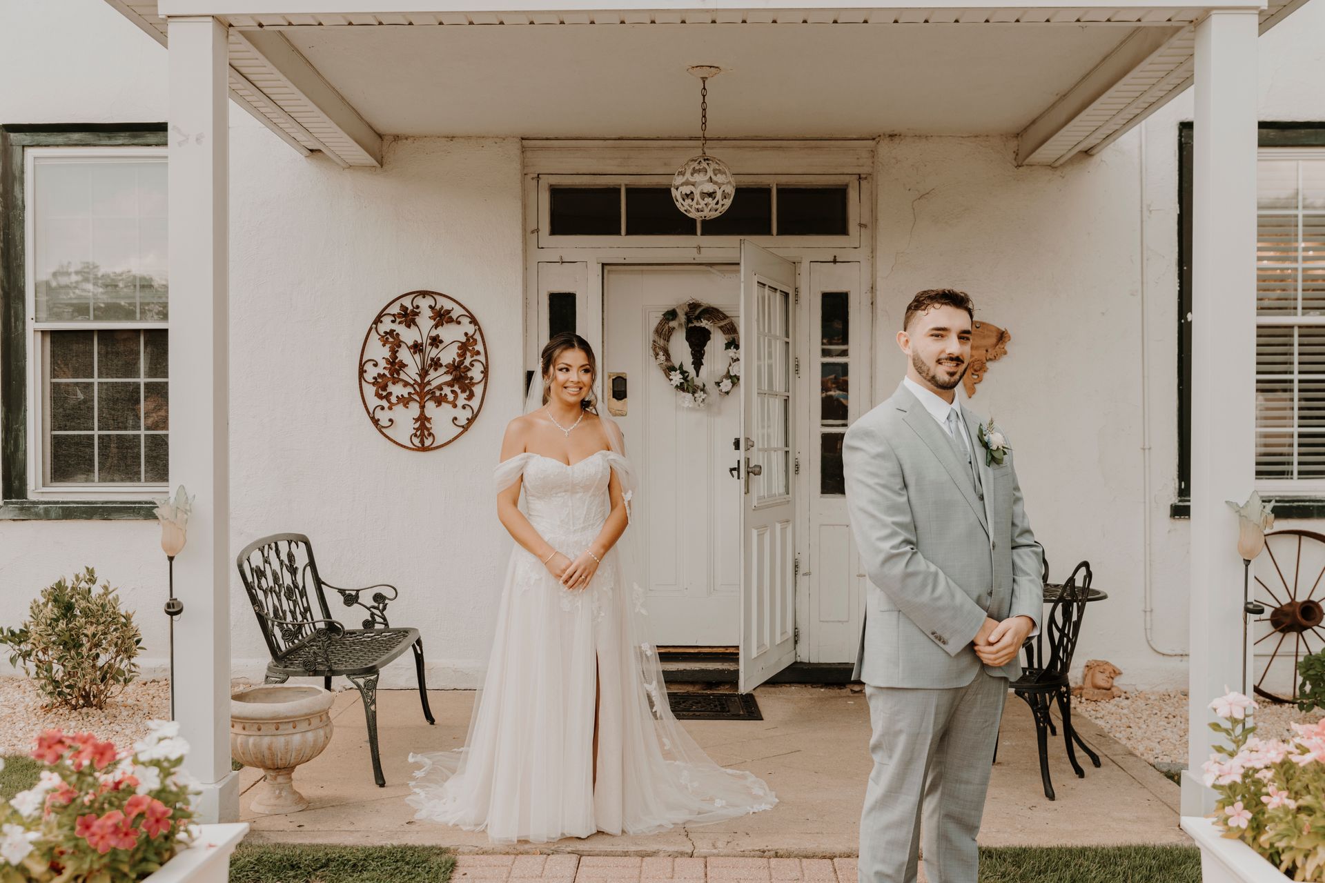 A bride and groom are standing on a porch in front of a white house.