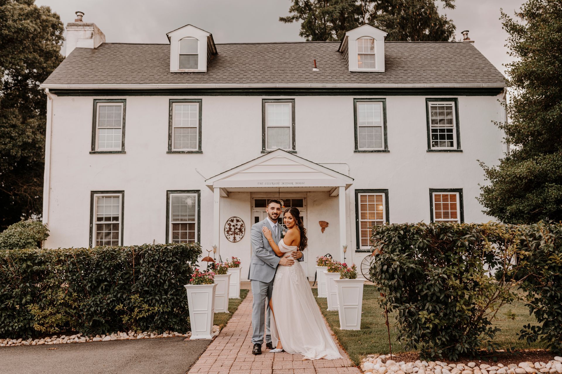 A bride and groom are posing for a picture in front of a large white house.