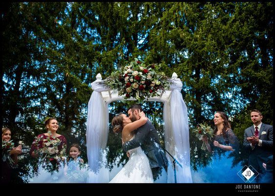 A bride and groom are kissing under an arch at their wedding ceremony.