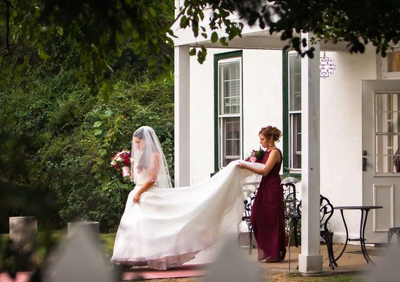 A bride and her bridesmaid are standing on a porch in front of a white house.