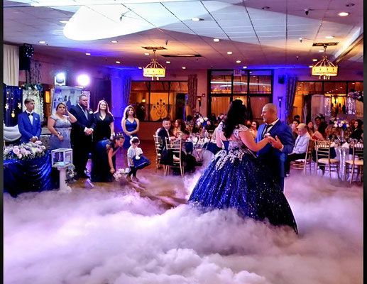 A bride and groom are dancing in a ballroom with smoke coming out of the ceiling.