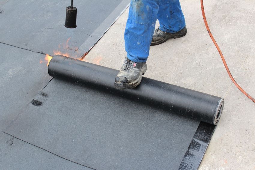 Person in blue overalls welding a black rolled roofing material onto a surface with a torch.