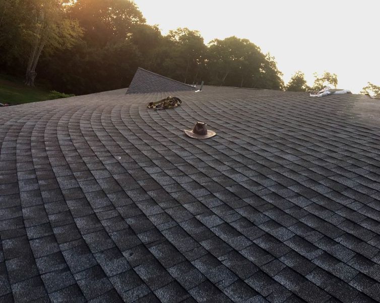 Dark gray shingled roof with a vent pipe, set against a backdrop of trees and a bright sky.
